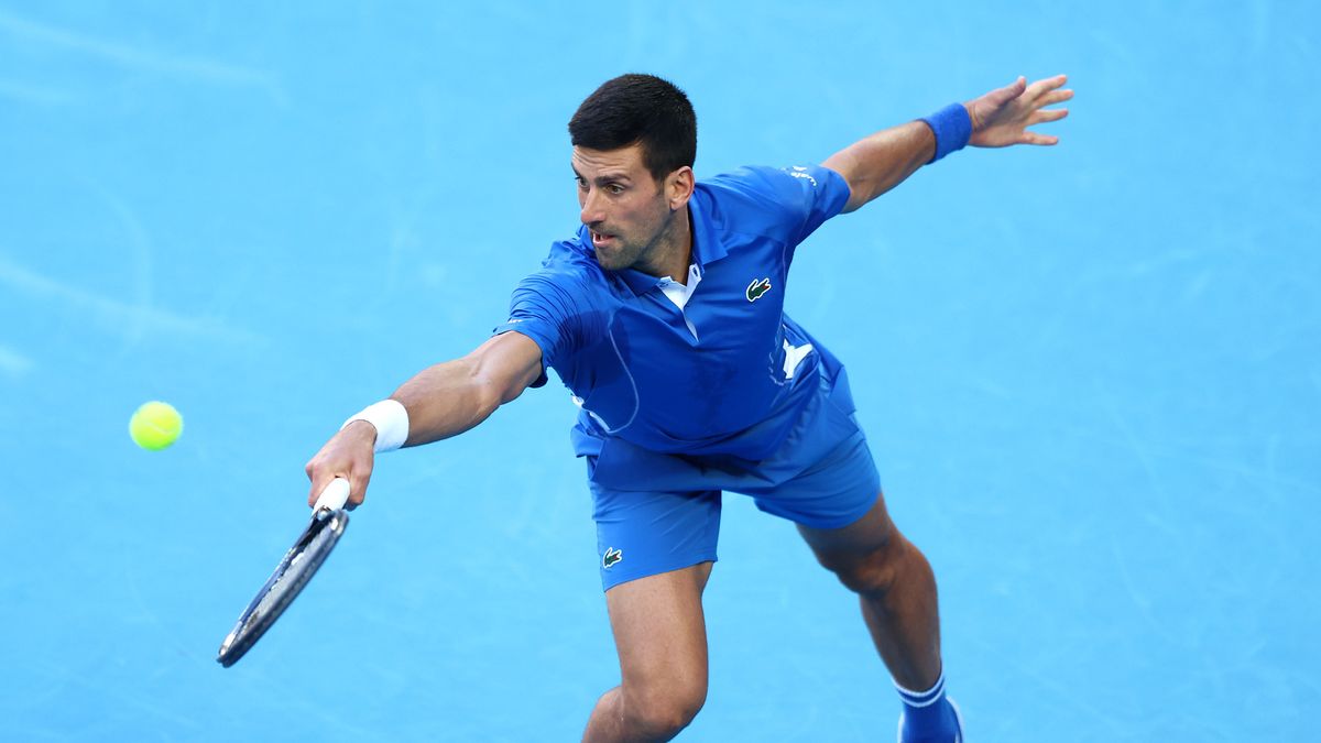 Novak Djokovic of Serbia, wearing royal blue tennis outfit, plays a lunging backhand on a blue court just prior to the Australian Open 2024