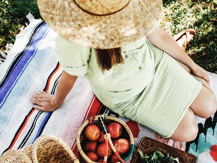 A pregnant woman sits on a blanket next to baskets of apples, nuts, and greens
