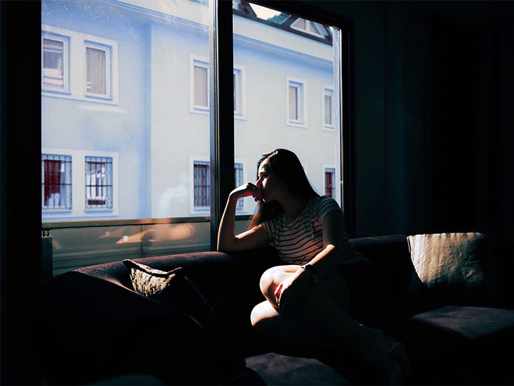 A woman looks out the window while sitting on a sofa in a dark room