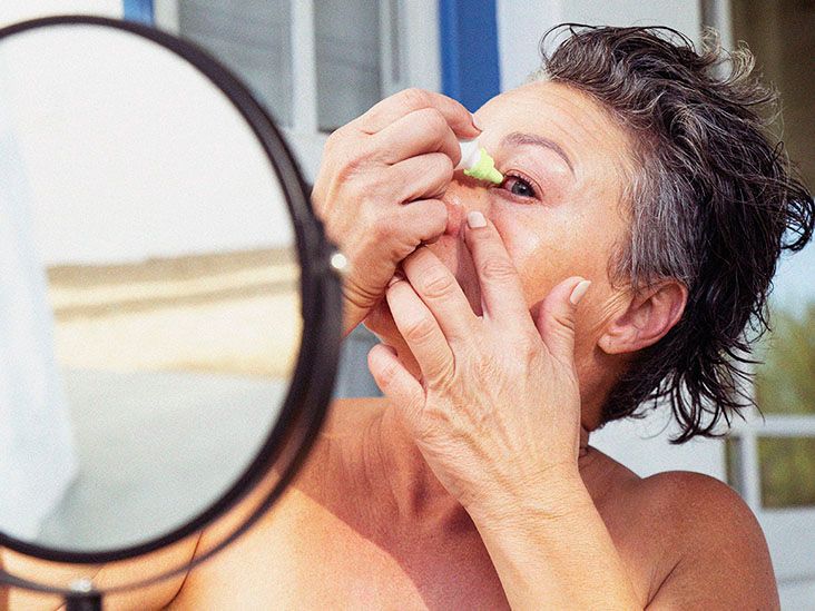 person with short black hair using eye drops in front of a round mirror