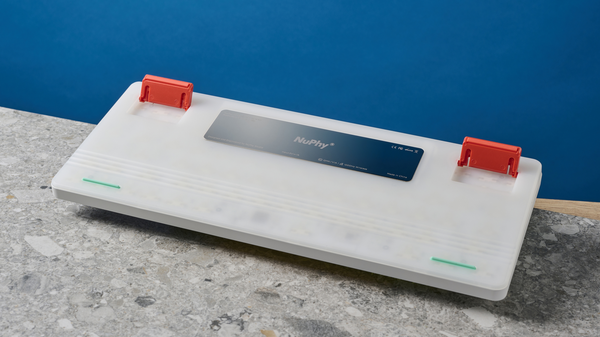תמונה של הצד התחתון של NuPhy Air75 V2 מראה את לוח המתכת והרגליים האדומות, על לוח אבן עם קיר כחול ברקע.