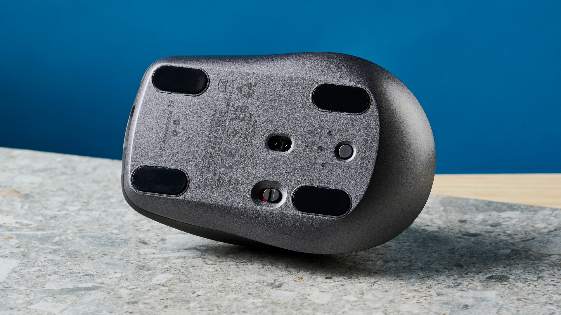 תמונה של עכבר Logitech MX Anywhere 3S הפוך על משטח אבן עם קיר כחול ברקע.