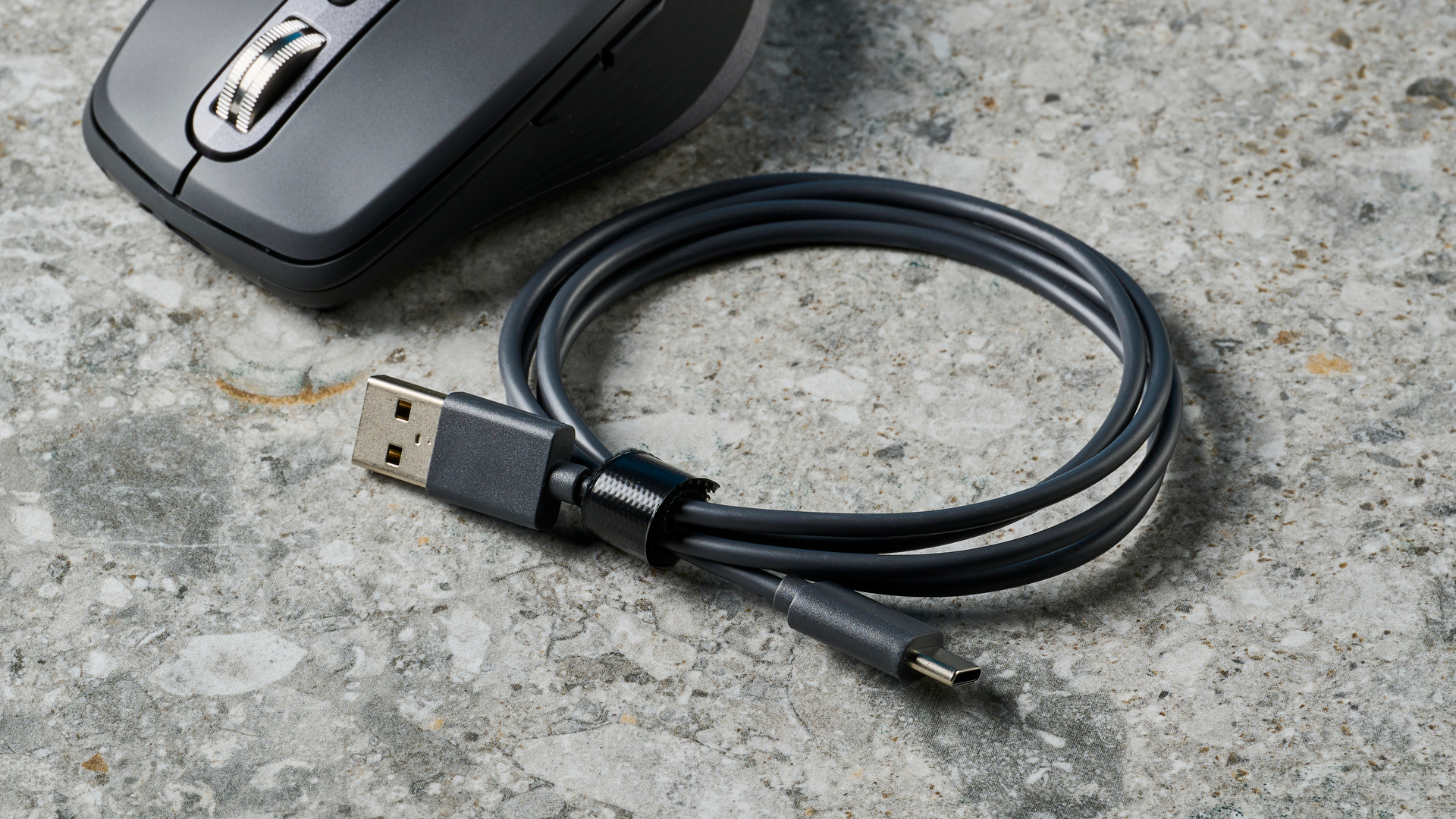 תמונה של עכבר Logitech MX Anywhere 3S על משטח אבן ולידו כבל USB-C ל-USB-A שלו.