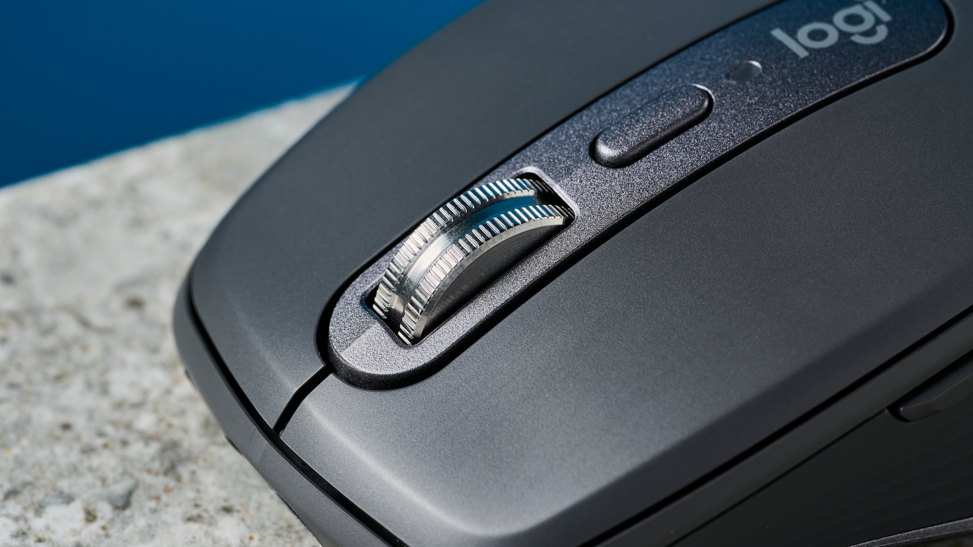 תמונה של עכבר Logitech MX Anywhere 3S על משטח אבן עם קיר כחול ברקע.