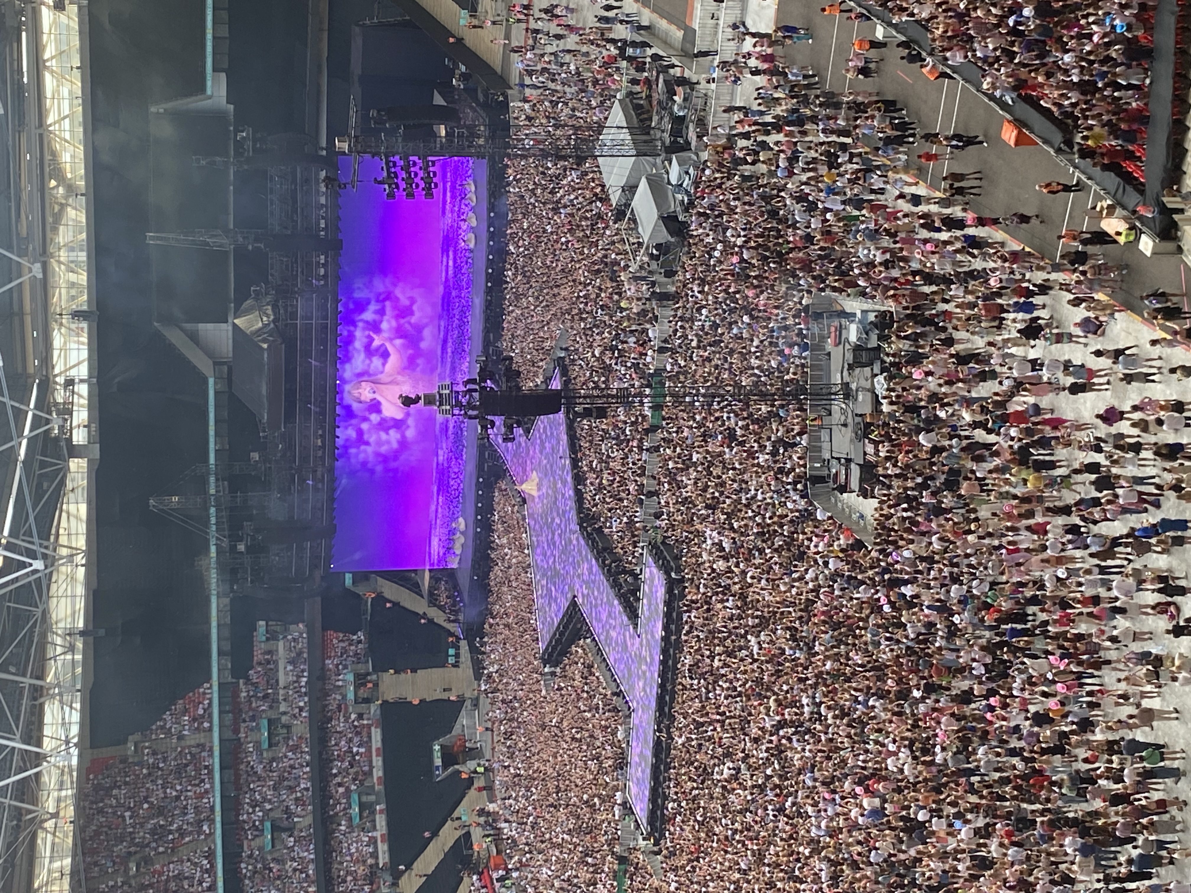 תמונה מ-The Eras Tour, לונדון בלילה 1. האצטדיון מלא וטיילור סוויפט נרתע על מסך גדול מאחורי שביל.  הנוף חסום מעט על ידי רמקול