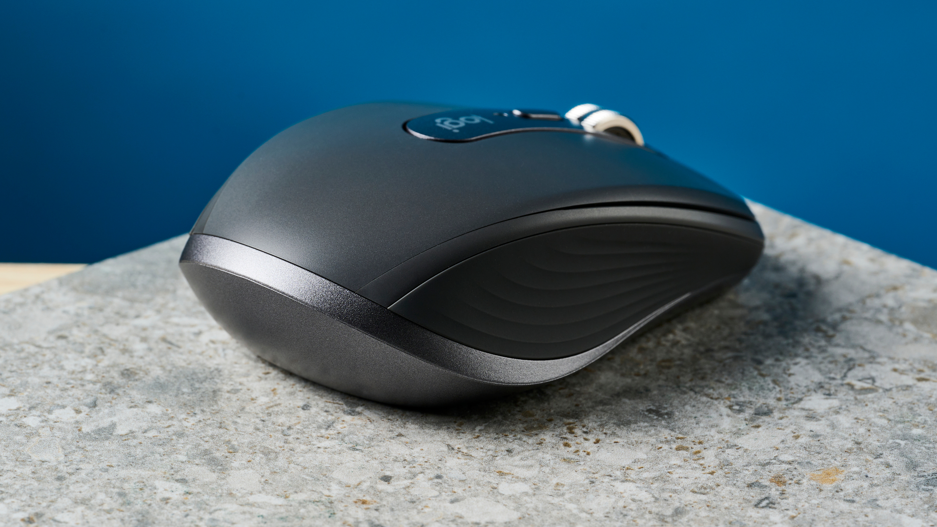 תמונה של עכבר Logitech MX Anywhere 3S על משטח אבן עם קיר כחול ברקע.