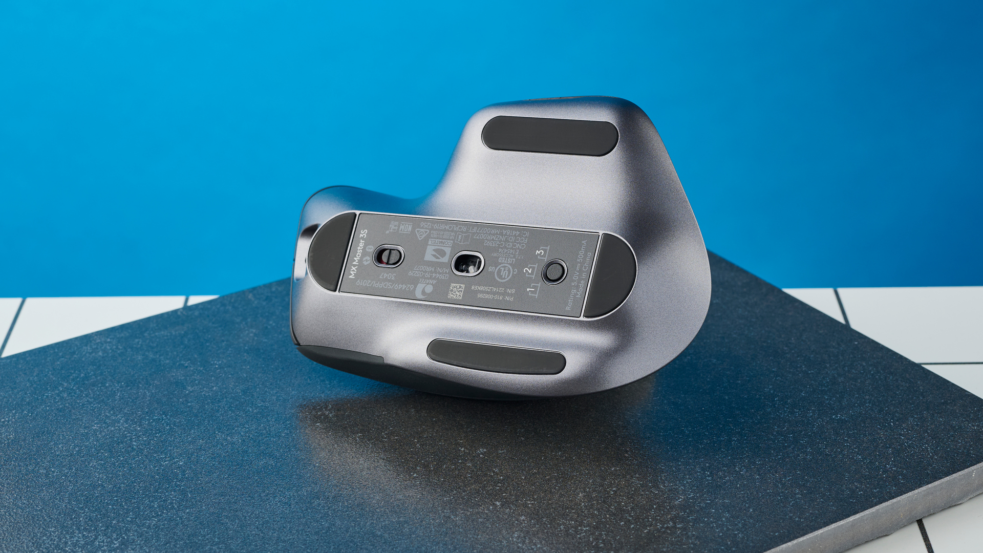 תמונה של Logitech MX Master 3S על צפחה שחורה על רקע כחול.  העכבר הפוך ומראה את החיישן האופטי.