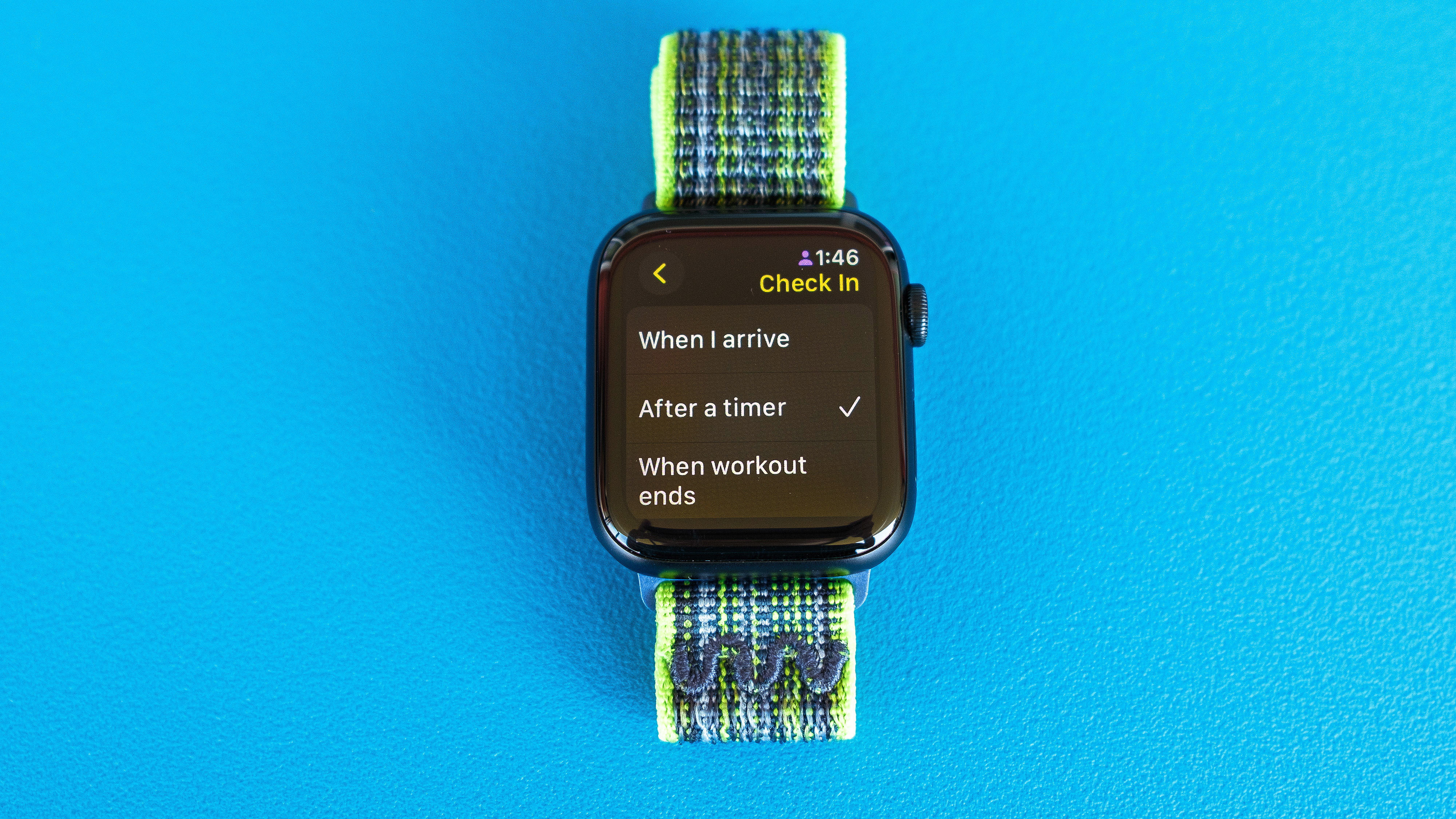 צילום פרט של Apple Watch SE על רקע כחול בהיר המציג את השלבים להגדרה ושליחה של הודעת צ'ק אין ב-watchOS 11 