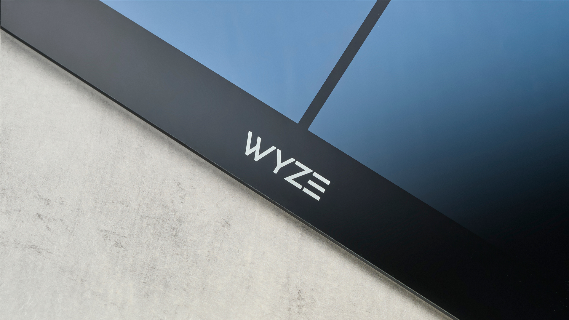 תמונה מקרוב של הלוגו של wyze בקנה מידה חכם מלוטש ונקי למראה עם גימור זכוכית מחוסמת ועיצוב בצורת צלב על גבי הזכוכית, כמעט כחול בתאורת הסטודיו, מונחת על שולחן