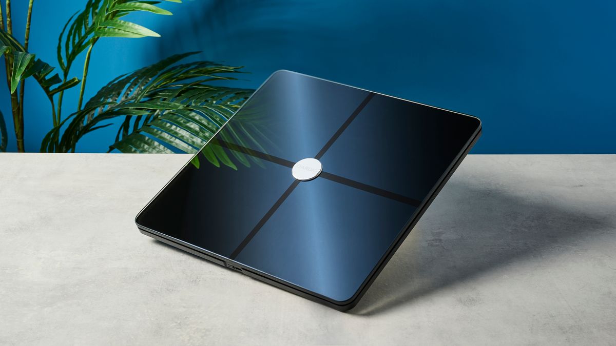 a modern black smart scale with a silver circle in its centre rests upon a light surface with a blue background