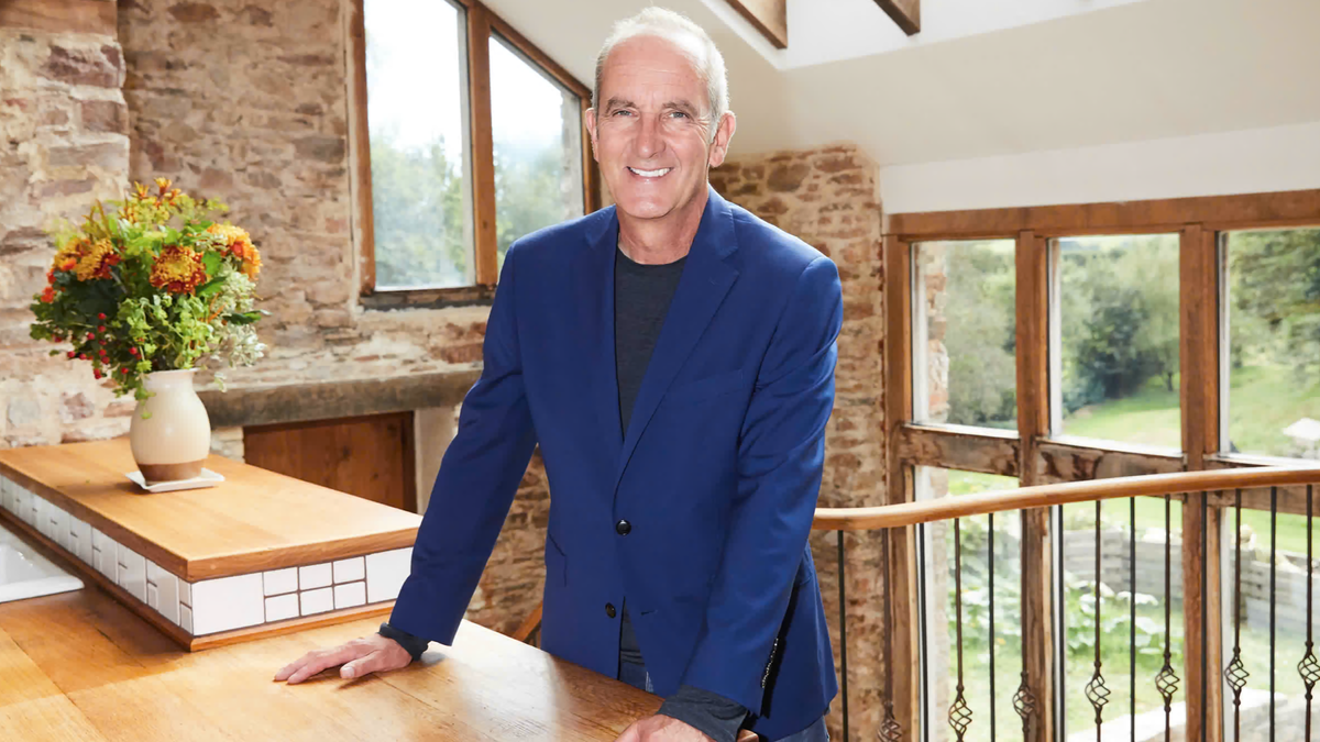Kevin McCloud, wearing a royal blue jacket, smiles in an airy but rustic cottage kitchen as he presents "Grand Designs: 25 Years and Counting"