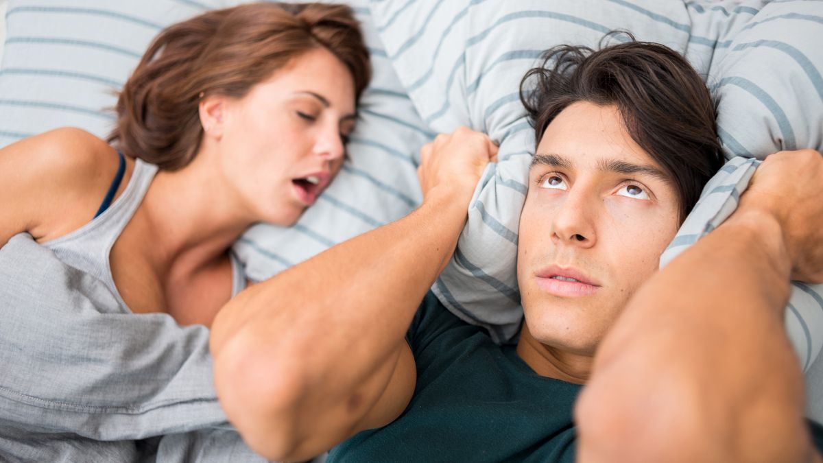 A man with dark hair rolls his eyes in frustration and holds a pillow to his ears because his partner is snoring. He is wondering how to stop snoring when sleeping