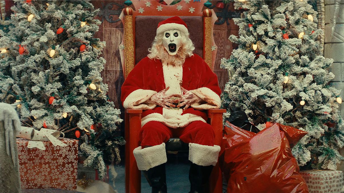 Art the Clown (David Howard Thornton) dressed as Santa Claus sitting on a throne flanked by Christmas trees and sacks of presents in "Terrifier 3"
