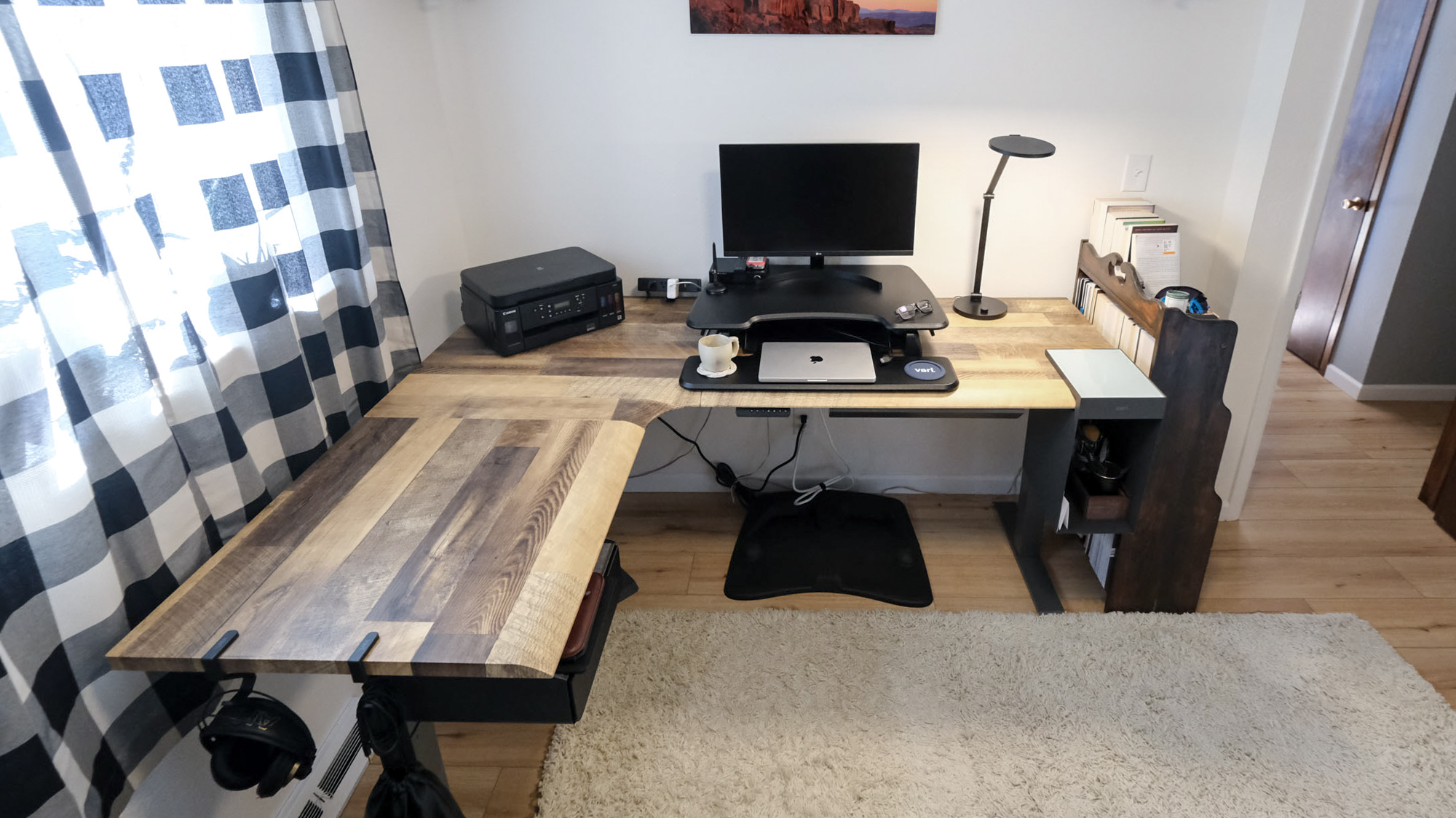 שולחן עמידה חשמלי Vari L-Shape בפינה של משרד ביתי עם מחשב נייד, צג, מדפסת ושאר אביזרי עבודה מהבית