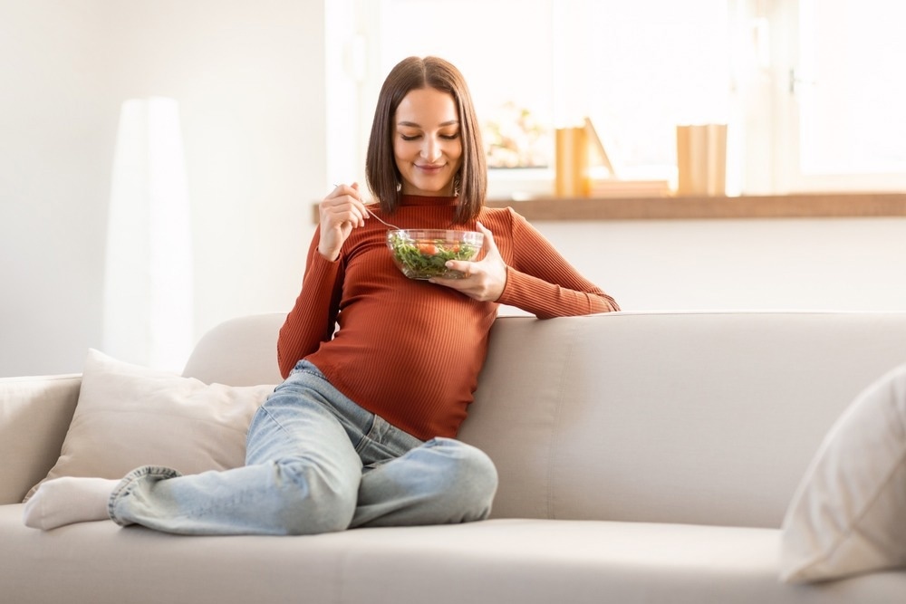 Pregnant woman enjoying a fresh vegetable salad while relaxing on a sofa indoors