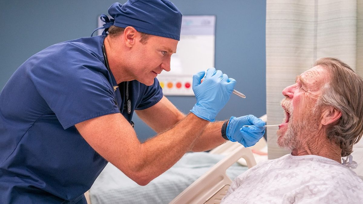 A doctor in blue scrubs comedically inspects a bed-bound patients' mouth in a scene from TV show "St. Denis Medical" season 1