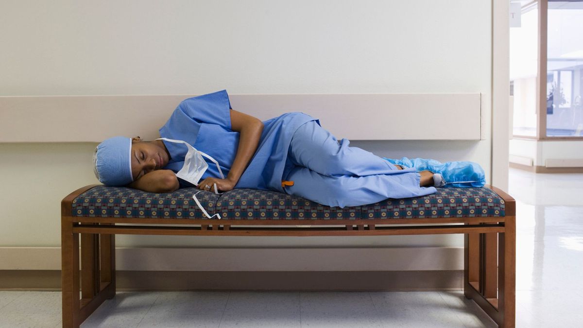 A nurse wearing blue scrubs asleep on her side on a bench in a hospital