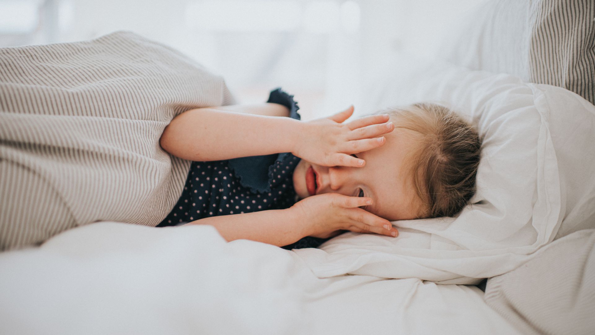 A child lying on their side covering their eyes in bed