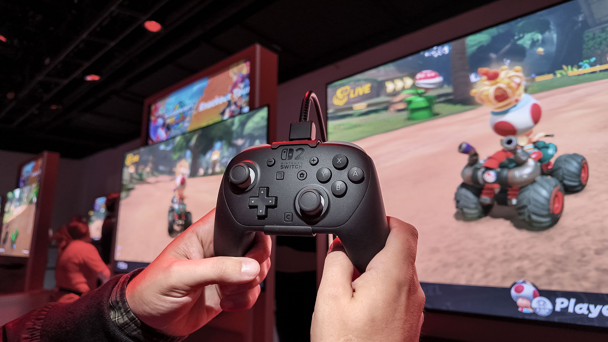 The Nintendo Switch 2 Pro Controller in hand with Mario Kart World on TVs in the background