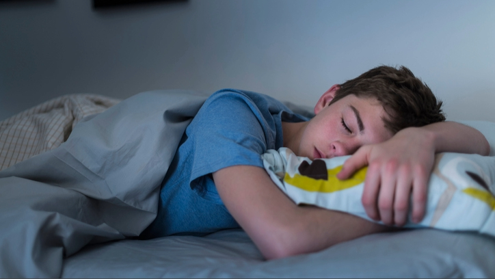 A teenage boy wearing a blue top sleeping on his front with one arm over head and one arm under pillow