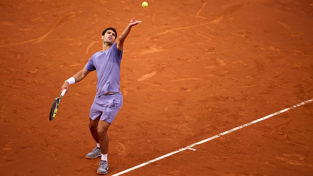 Carlos Alcaraz, dressed in lilac, serving on a clay court at Real Club De Tenis Barcelona