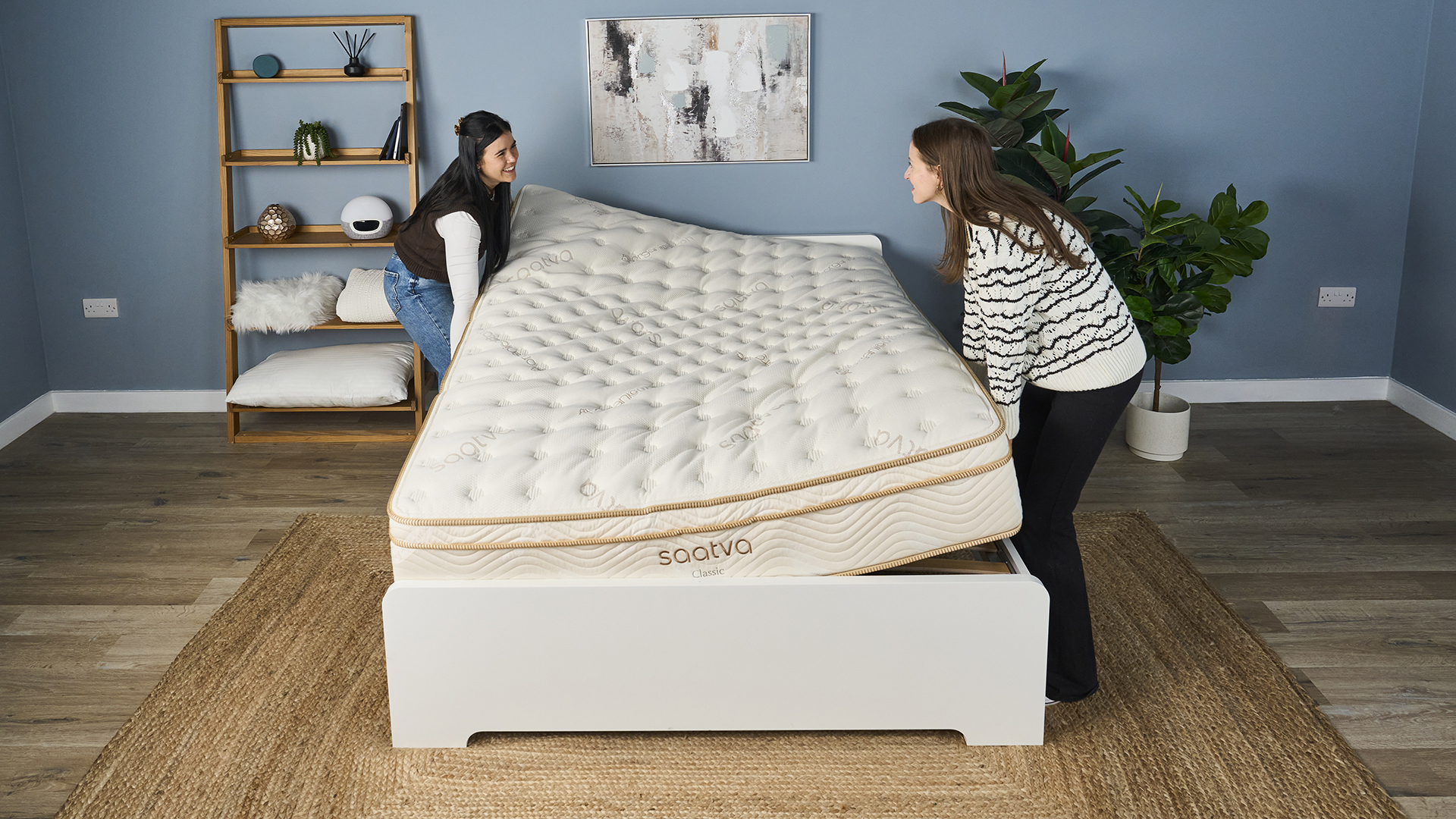 Two women rotate the Saatva Classic mattress onto a bed frame in a bedroom.