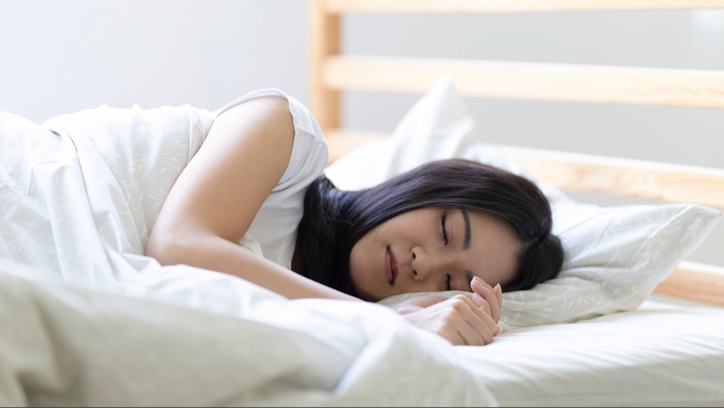 A woman with dark hair sleeping on her side in bed with white bedding