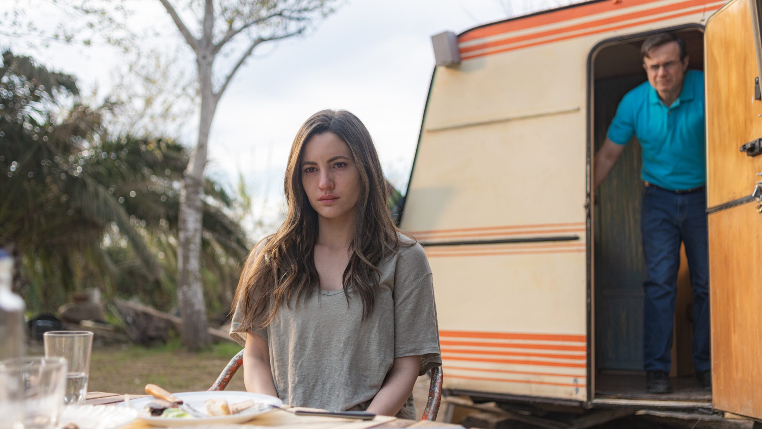 Maje (Ivana Baquero) sits in front of a caravan with Salva (Tristán Ulloa) in the doorway behind her in a scene from Netflix's "A Widow's Game"