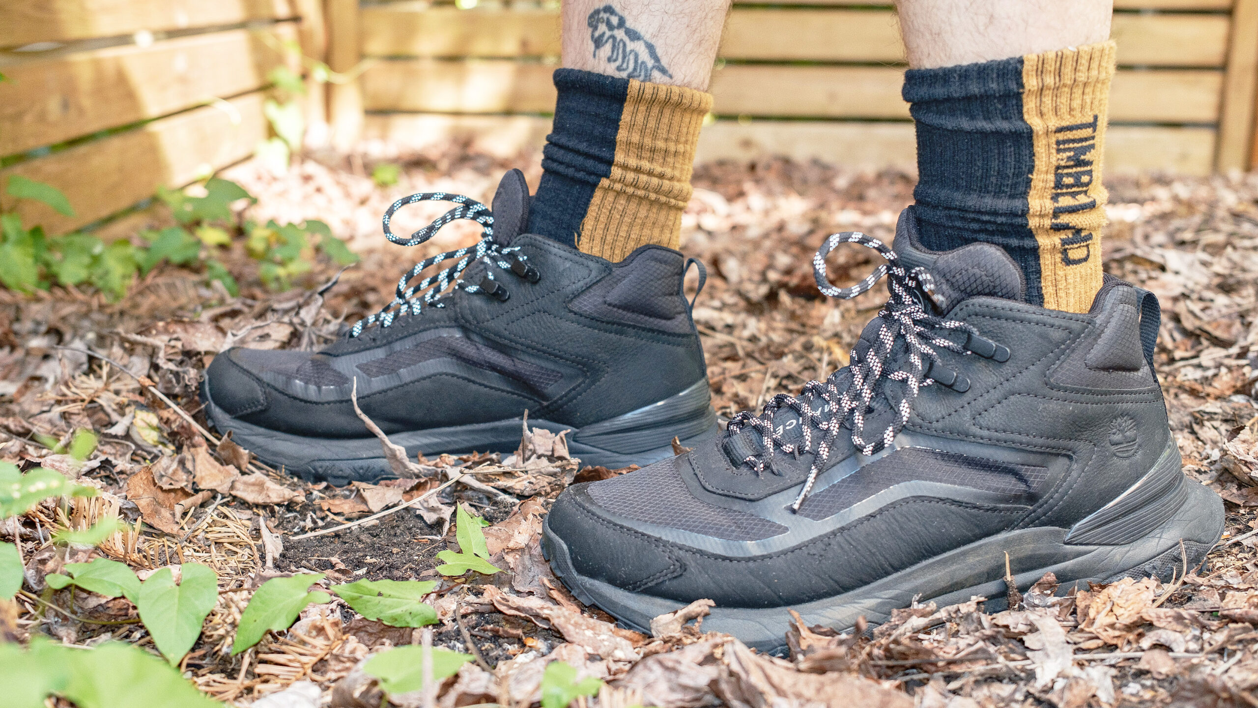 Close-up of the side of the Timberland Motion Access Mid Waterproof Sneakers (shown in black) on a hiker's feet shown standing in leaves