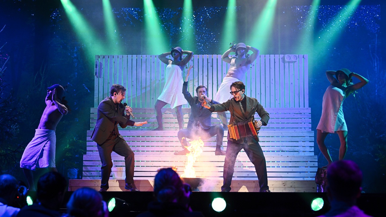 Swedish music group KAJ perform their entry "Bara bada batsu" on stage, in front of a mock recreation of a sauna, while flanked by backing dancers wearing white towels and bucket hats.