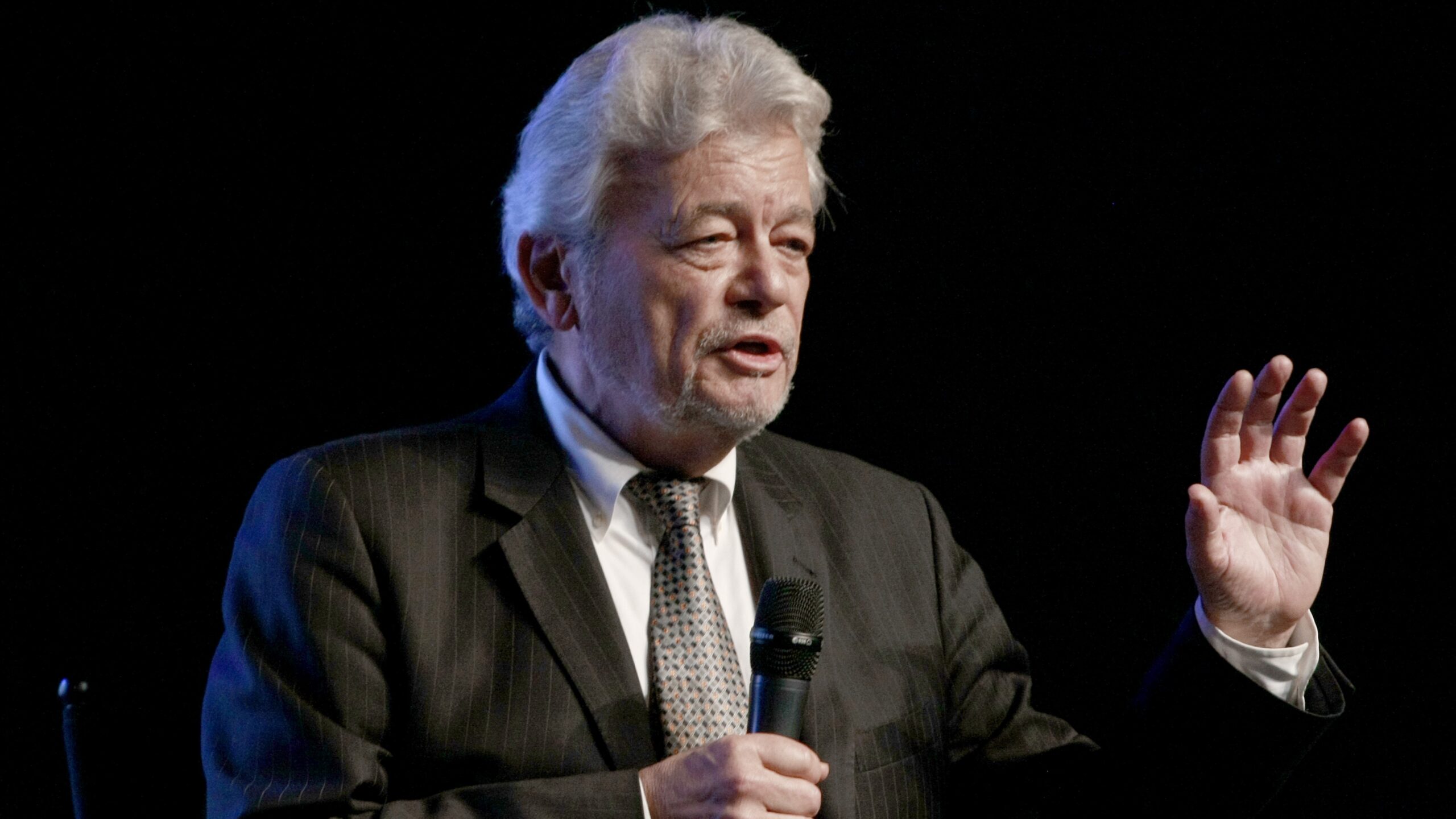 George Knapp, wearing a dark suit and tie, speaks onstage at HISTORY's Unidentified panel during AlienCon Los Angeles 2019 ahead of Contact in the Desert 2025 live stream of the UFO conference in California.