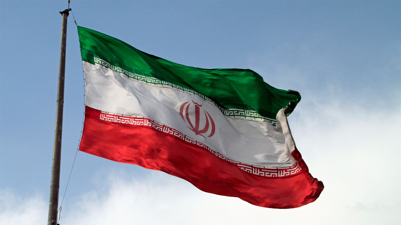 Flag of Iran flying in front of cloudy blue sky. The flag has three horizontal stripes, coloured green, white, and red, with a red symbol in the middle