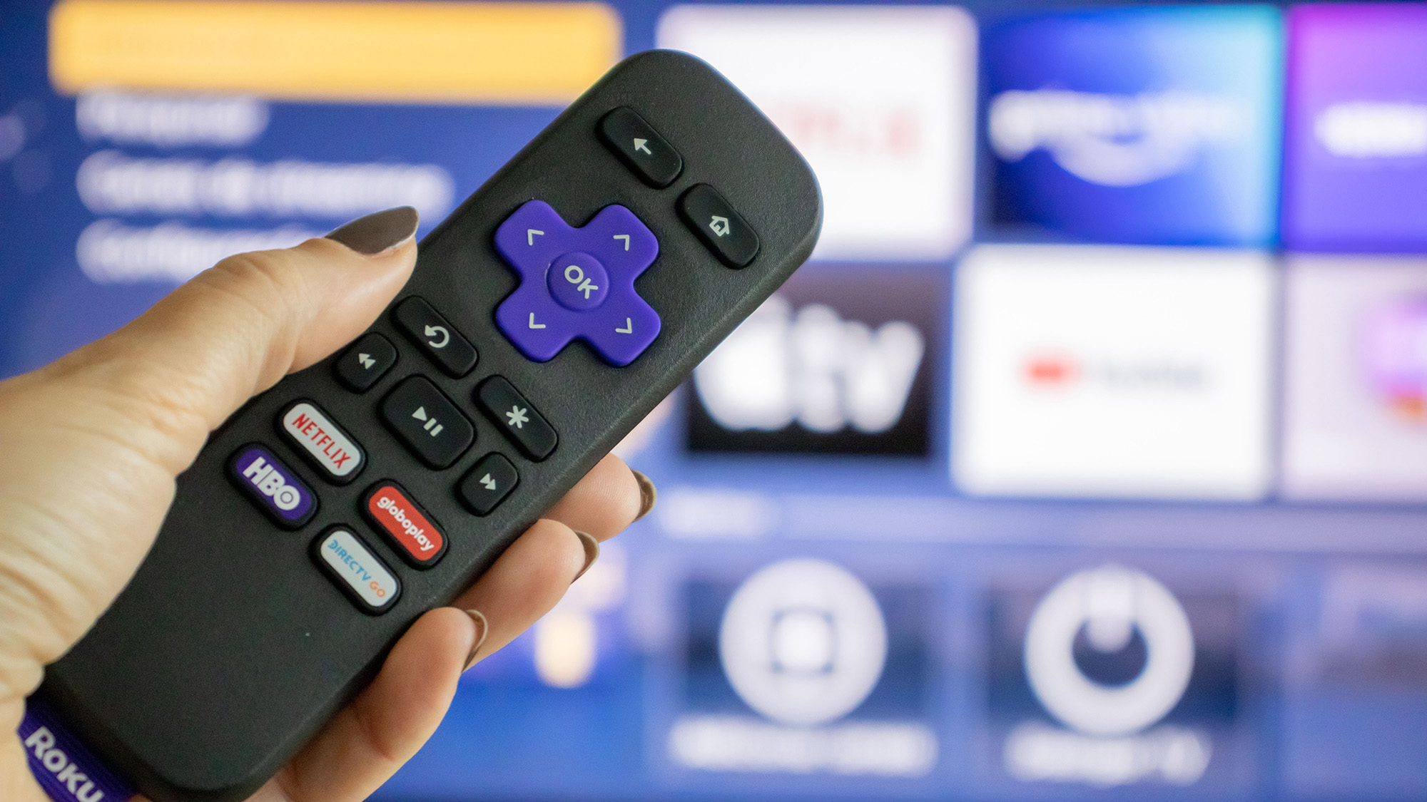 יד מחזיקה שלט של Roku מול טלוויזיה עם מסך הבית של Roku.