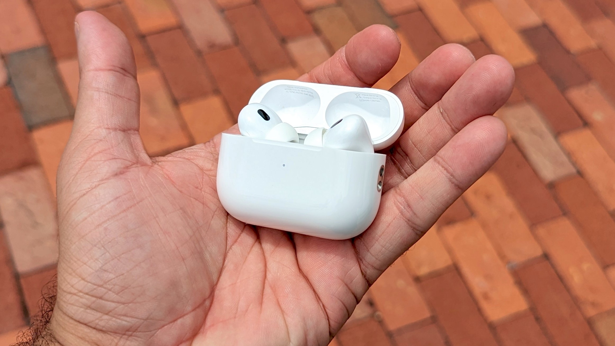 AirPods Pro 2 בתיק טעינה ביד הסוקרת עם ריצוף לבנים אדומות ברקע