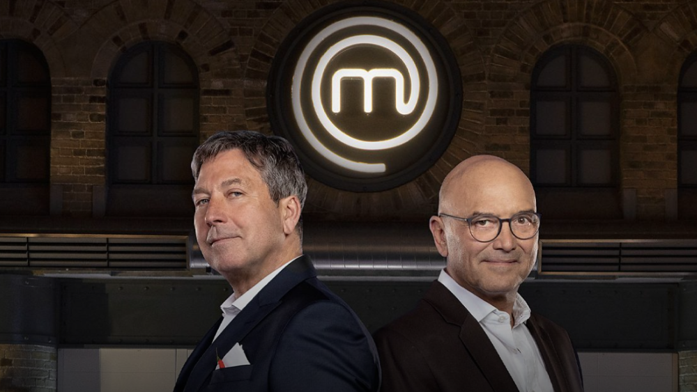 Presenters John Torode and Gregg Wallace standing beneath the MasterChef UK crest