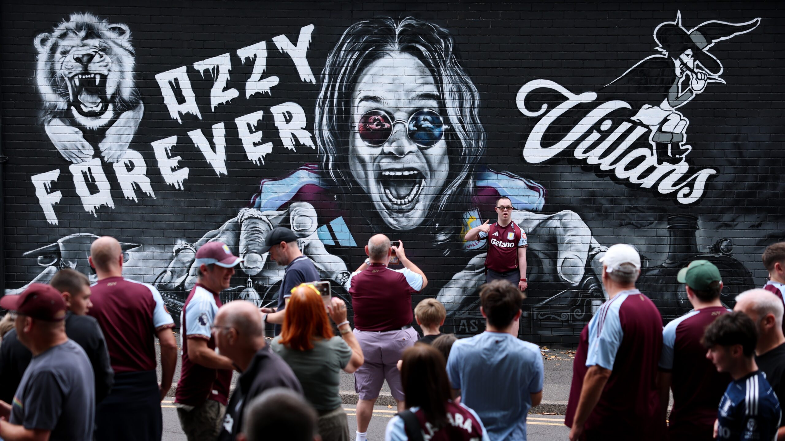 Fans pose for a photo in front of an Ozzy Osbourne mural prior to during the Premier League match between Aston Villa and Newcastle United at Villa Park on August 16, 2025 in Birmingham, England.