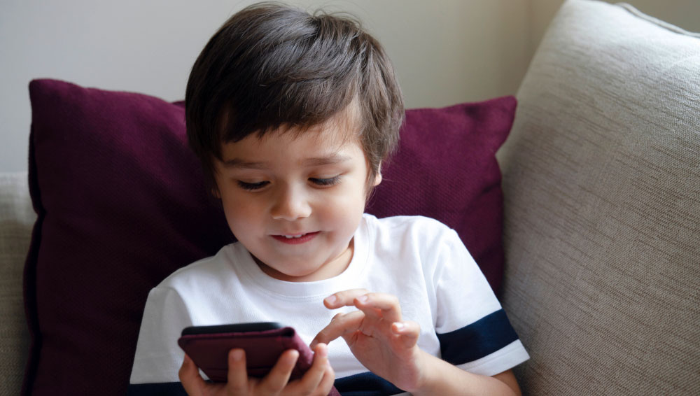 ילד כהה שיער בערך 5 באמצעות Smarthphone בזמן שהוא יושב על הספה.