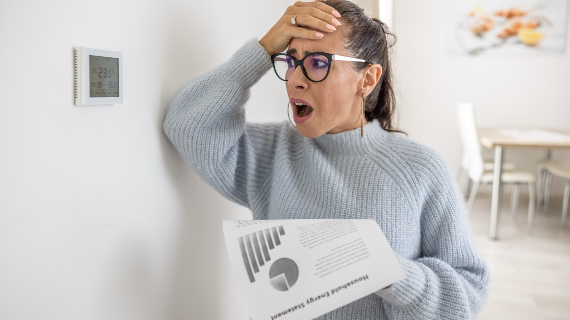 woman holding energy bill next to thermostat looking upset