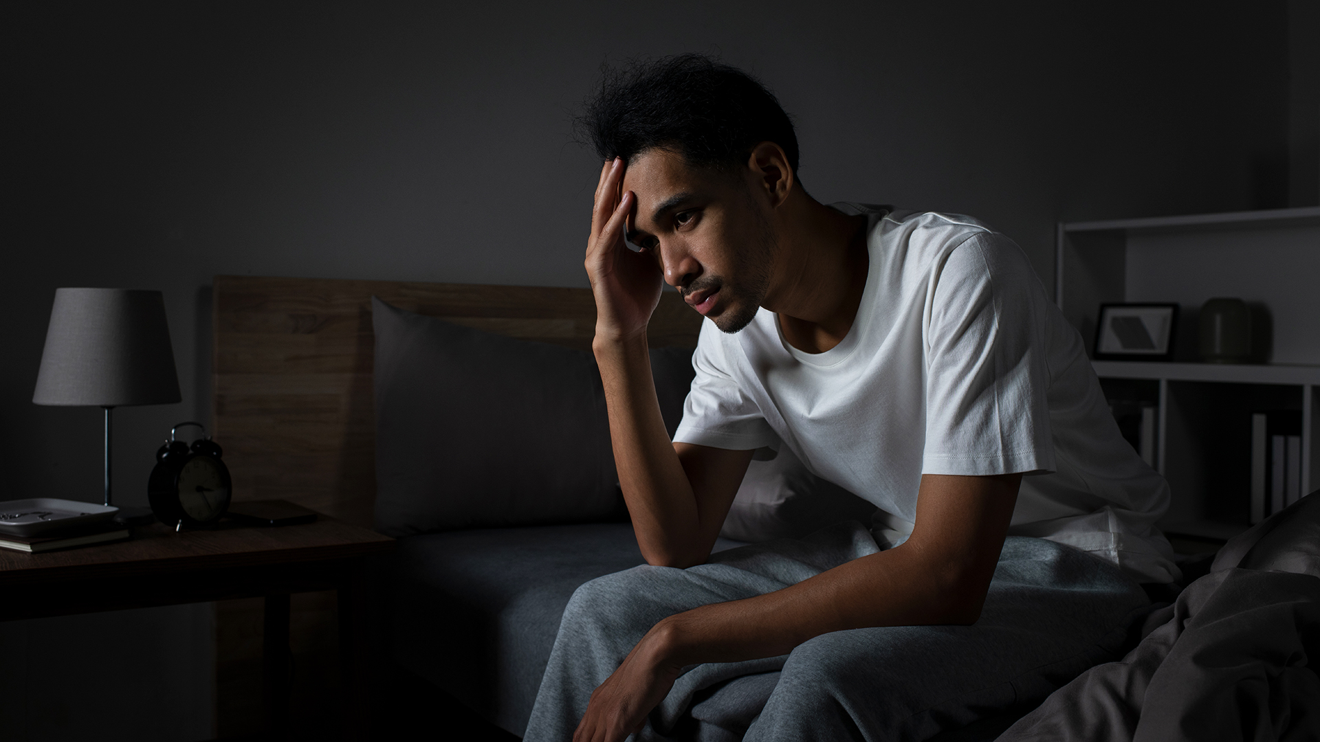 גבר יושב בצד מיטתו בחדר חשוך עם ראשו בידיו, כי הוא חווה מצוקה מחוסר יכולת לישון