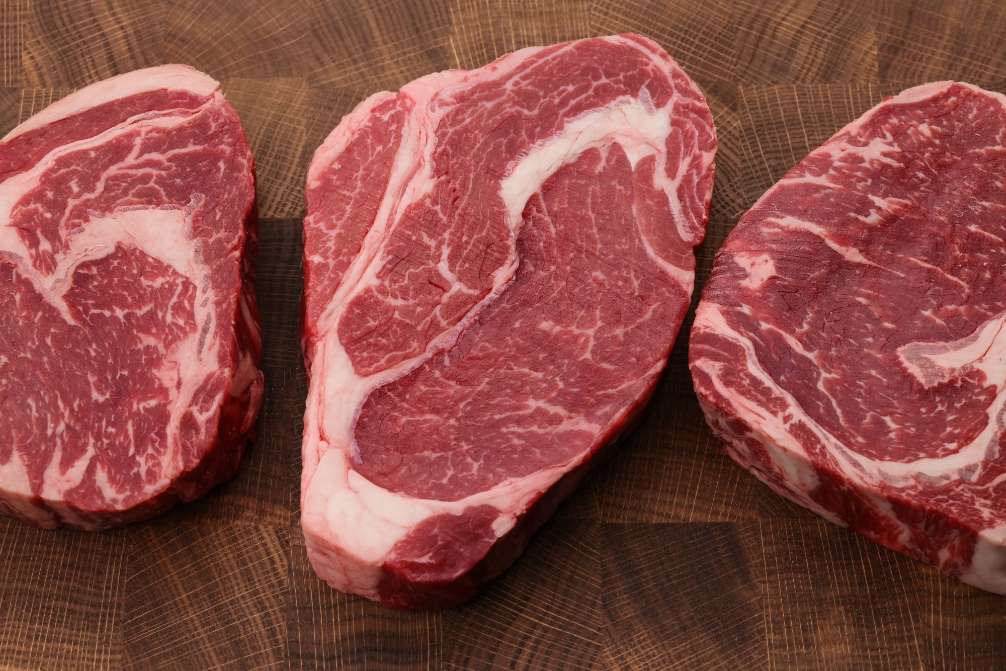 Close up three raw marbled ribeye beef steaks over end grain cutting board of wooden butcher block, high angle view, directly above.