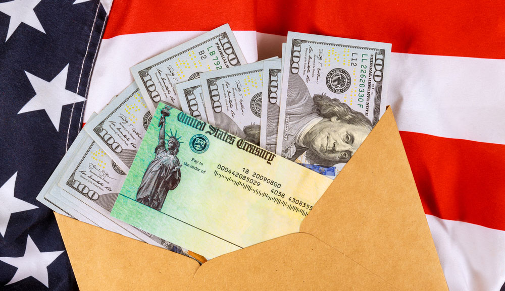 איור של שטרות של מאה דולר וצ'ק ממשרד האוצר האמריקני מבצבץ מתוך מעטפה של מנילה על רקע הדגל האמריקאי.
