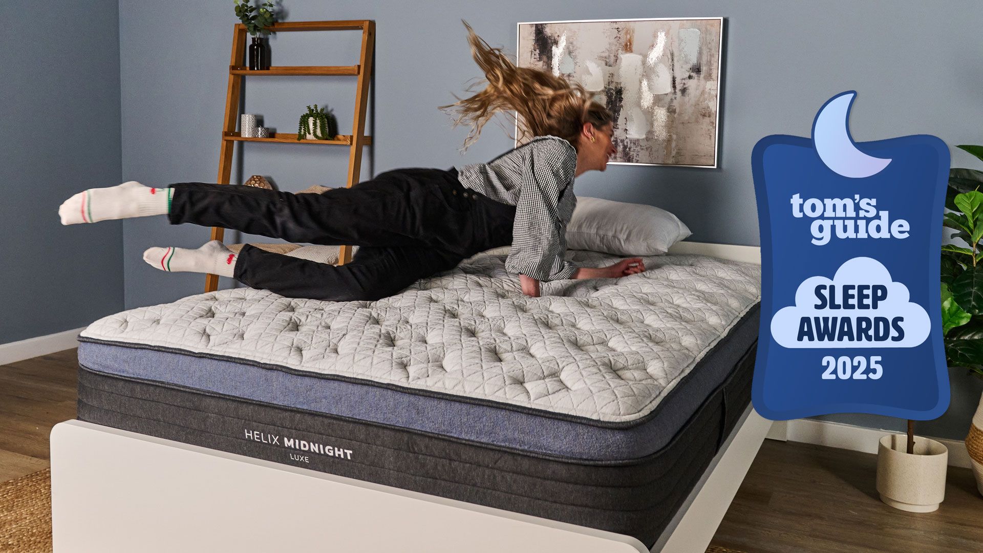 A woman diving onto a white pillow top mattress with blue trim, on a white bed frame in a blue bedroom, with a Tom's Guide Sleep Awards badge overlaid