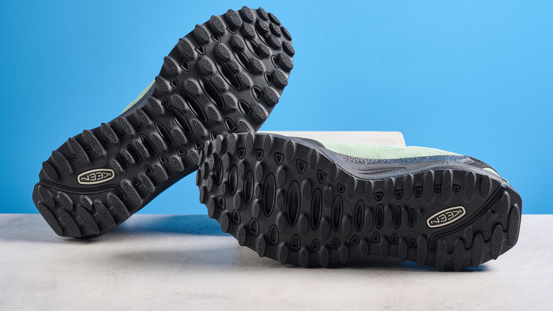 נעלי ההליכה Keen Jasper Zionic בצבע ירוק/זהב מצולמות על רקע כחול של Tom's Guide ורצפת אבן, מציגות את סוליית הזיז ואת החלק העליון של העור עם שרוכים כתומים