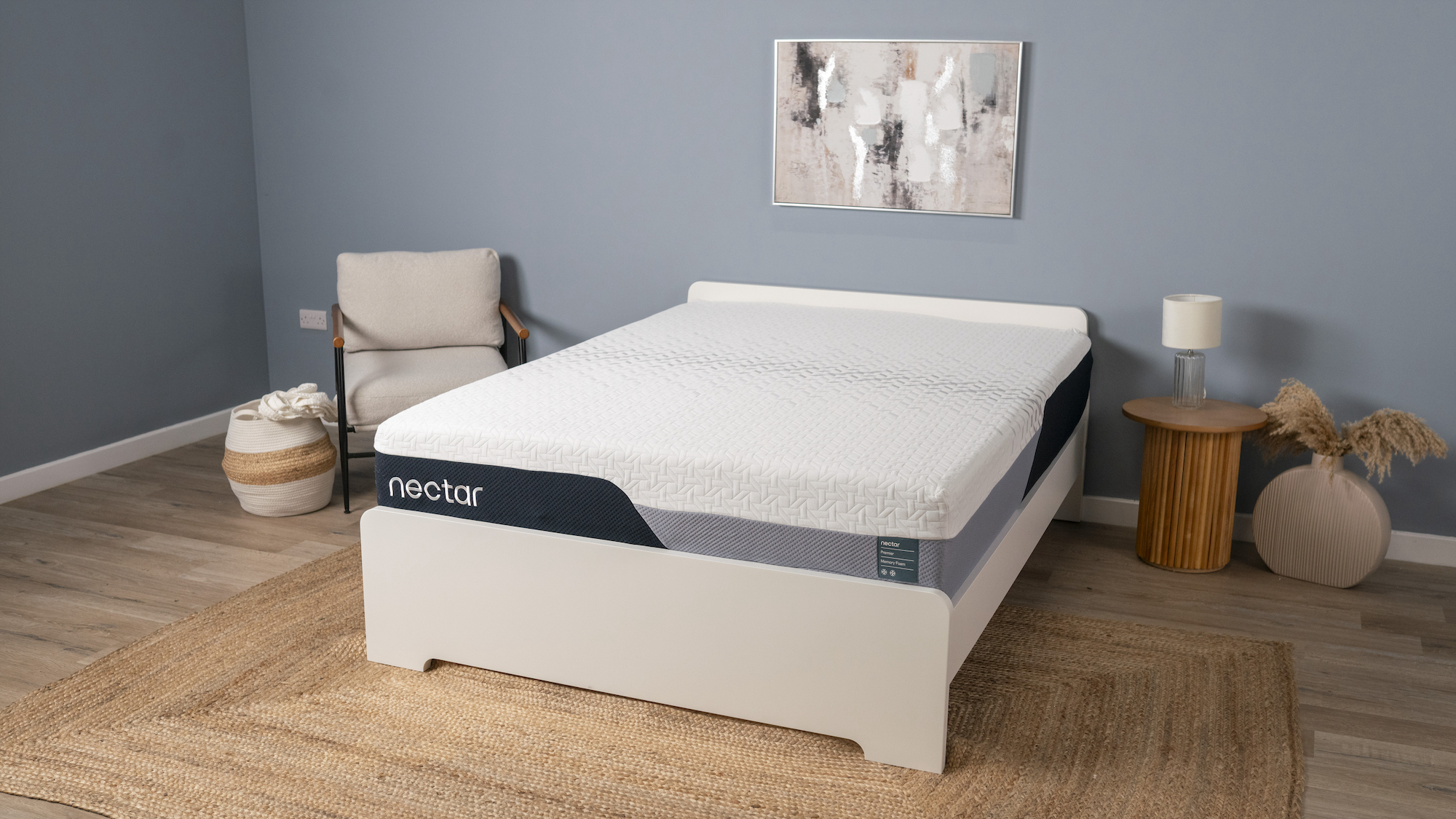 מזרון ה-Nectar Premier Memory Foam צולם על מסגרת מיטה לבנה מעץ במעבדת השינה הכחולה שלנו, עם כורסת בד בז' מסוגננת לצד המיטה ומתחתיה מחצלת קש גדולה