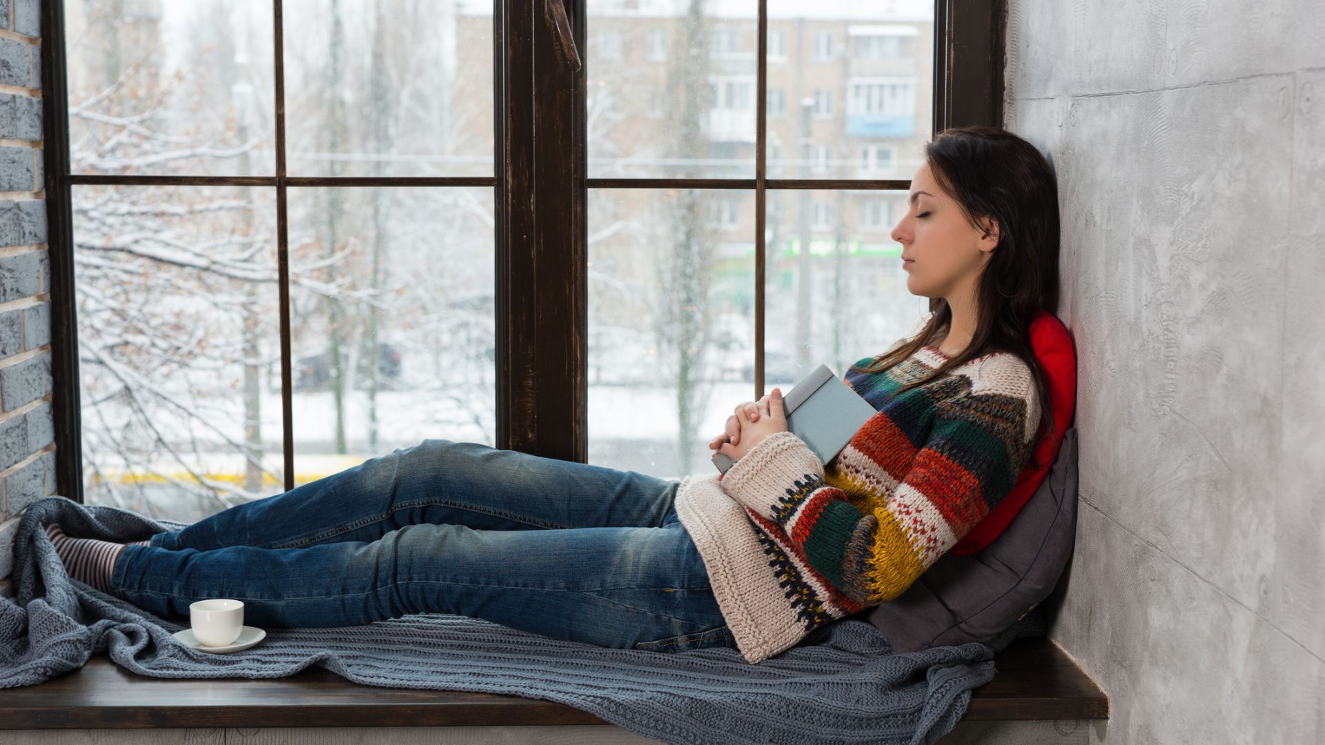 A woman sleeping in a windowsill wearing a knitted jumper with snow falling outside
