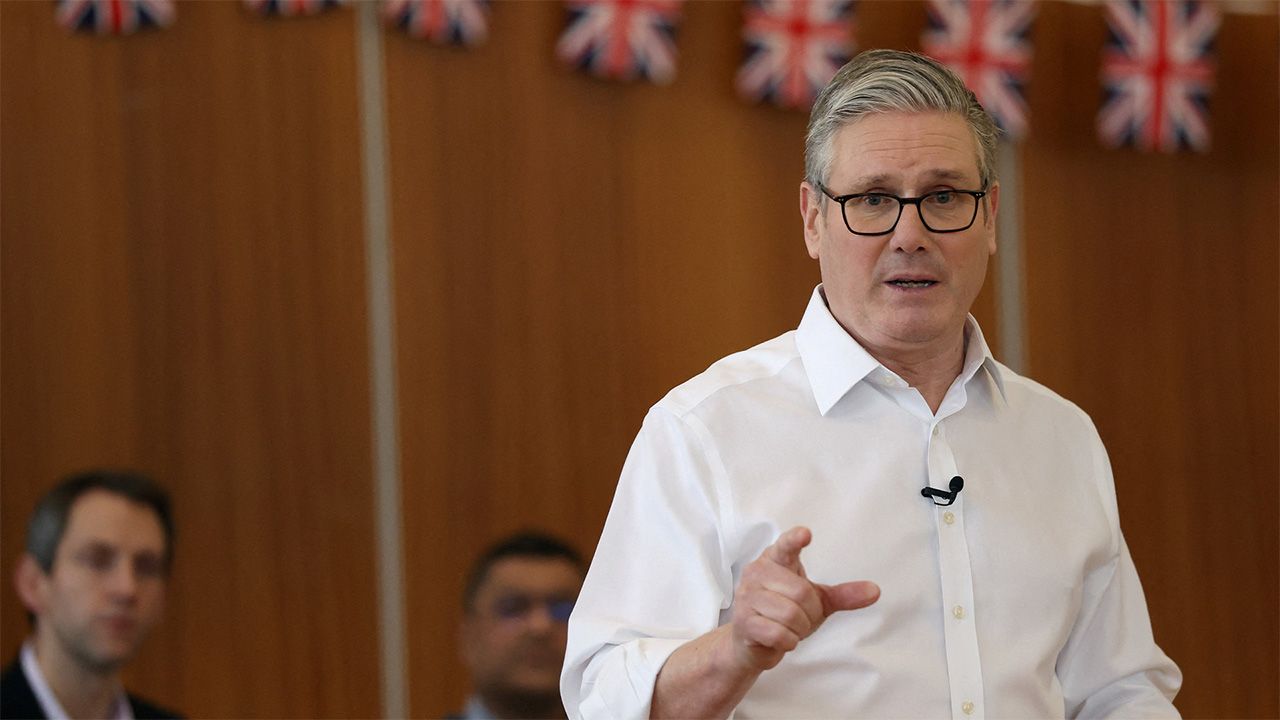 UK Prime Minister Keir Starmer delivers a speech in front of brown wall draped in Union Flags.