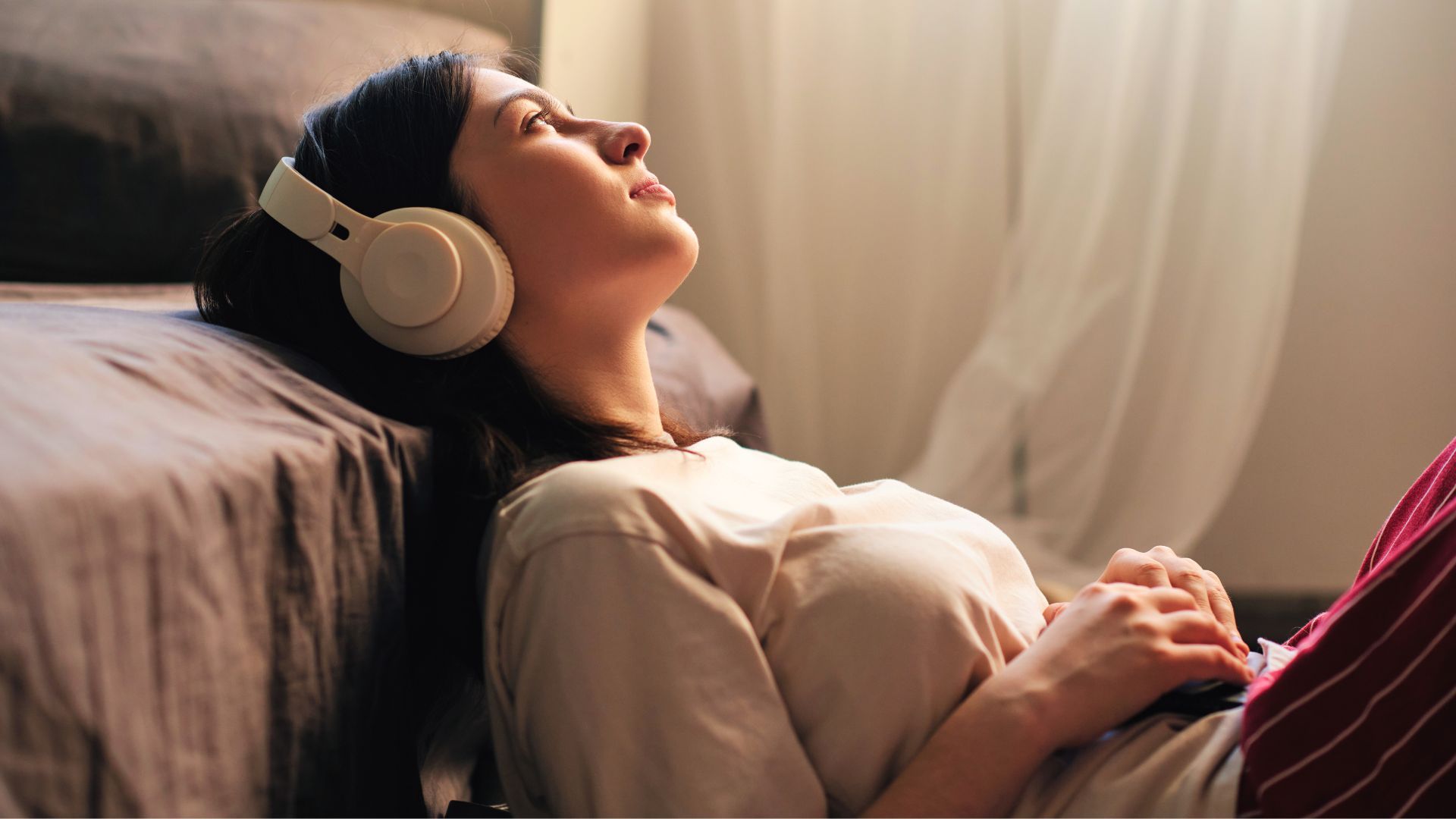 A young brunette woman relaxes against her bed while listening to a podcast through headphones.
