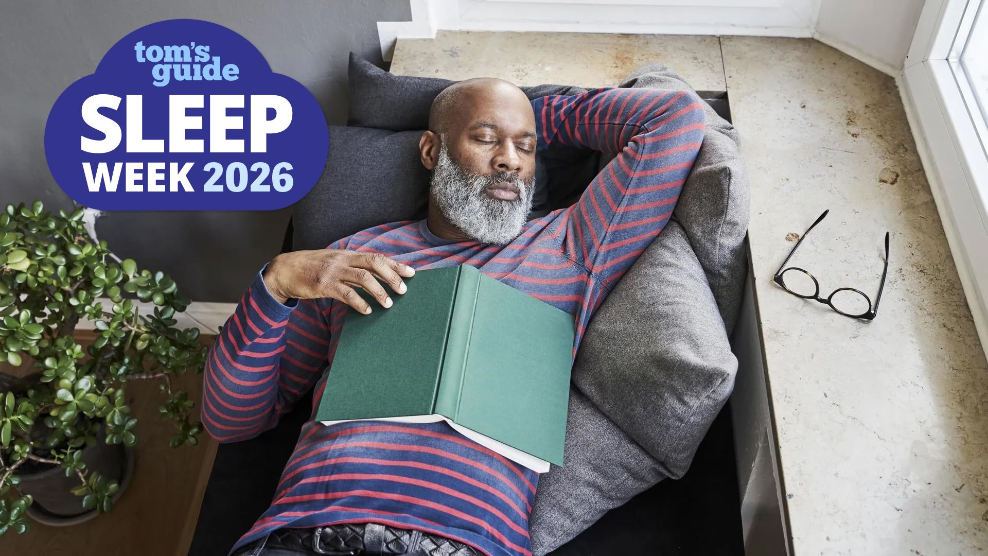 A man in his late 60s, dressed in a striped red and blue sweatshirt, takes a nap on the couch after reading a book. He has no idea that his long daytime naps as the reason why he keeps waking up at 3 a.m. A blue Tom's Guide sleep week logo sits in the top left hand corner.