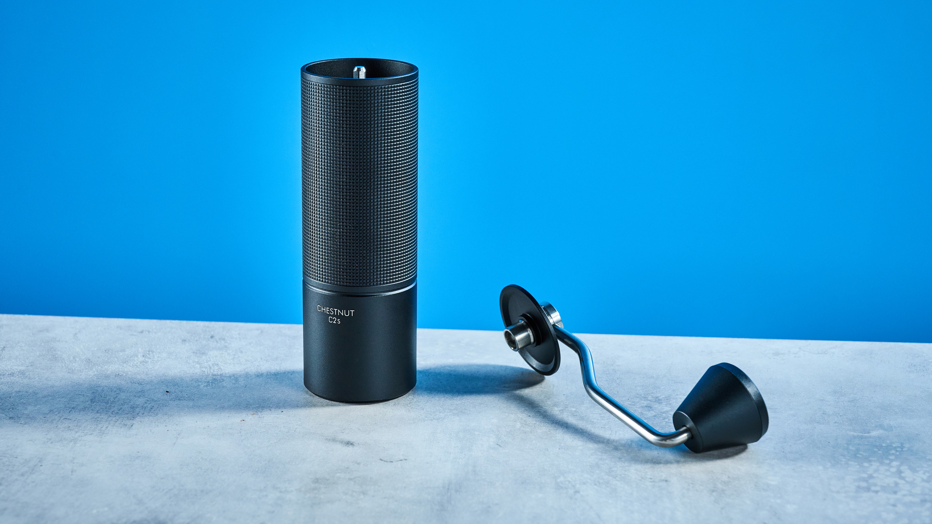 מטחנת הקפה של timemore chestnut c2s בשחור, מציגה את ארכובה היד ואת תא התפיסה שלה שצולמו על רקע המדריך הכחול של Tom's