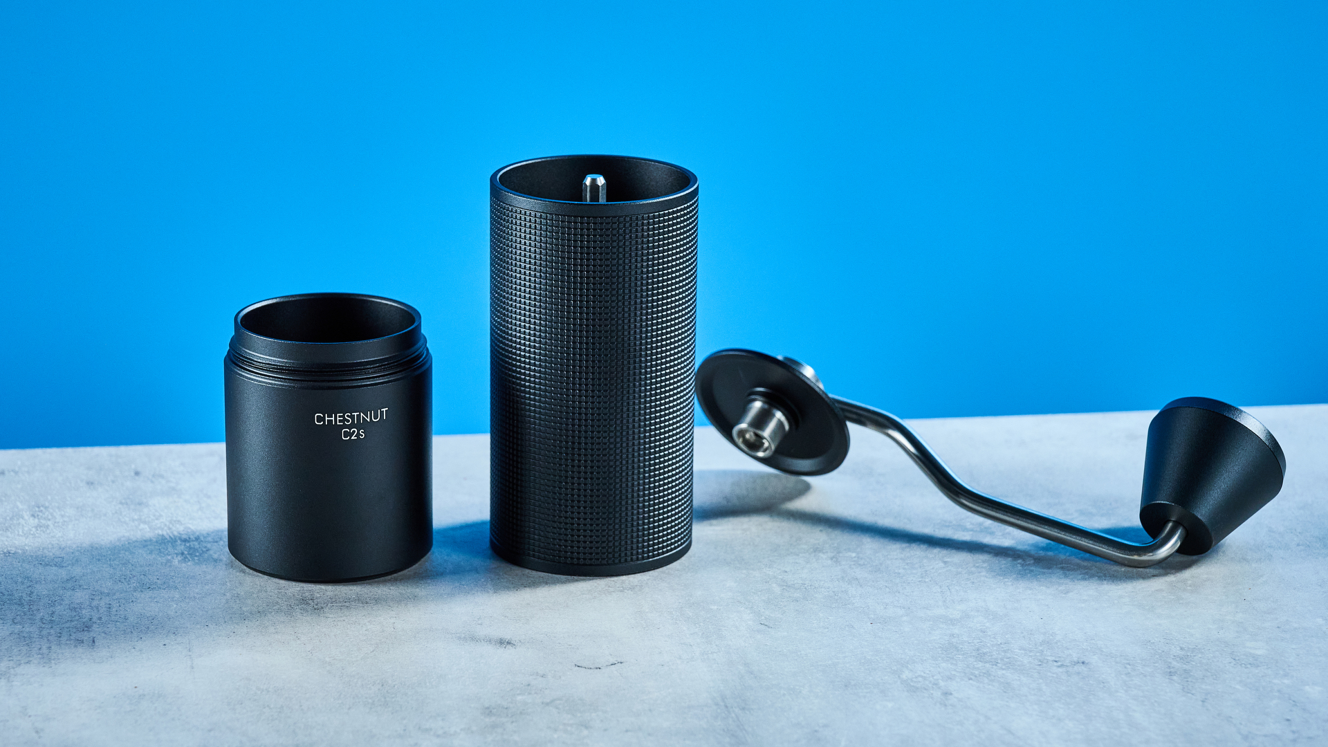 מטחנת הקפה של timemore chestnut c2s בשחור, מציגה את ארכובה היד ואת תא התפיסה שלה שצולמו על רקע המדריך הכחול של Tom's